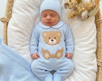 May include: A light blue knitted footed sleeper with a white Peter Pan collar and a central tan teddy bear appliqué wearing a blue and white checkered shirt. The outfit includes a matching light blue knit beanie with a small pom-pom on top.