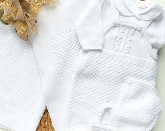 May include: A white knitted baby outfit set consisting of a long-sleeved shirt with a rounded collar, a pair of footed overalls with a textured diamond pattern and button details, and a matching bonnet, accompanied by a white knitted blanket.