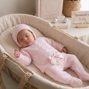 Könnte beinhalten: Ein schlafendes Baby trägt ein rosa Strick-Outfit, bestehend aus Mütze, Pullover und Hose, in einer gewebten Babywiege. Das Outfit hat eine Schleife und dekorative Spitze. Ein Schild liest "Nacimiento de Baby Bienvenida al Mundo."