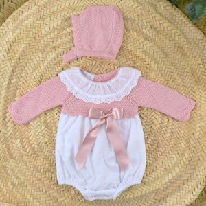 May include: A pink knitted baby romper with long sleeves, a white dotted front, and a white lace collar. A matching pink bonnet is also included. The romper features a satin bow at the waist, and the set is displayed on a woven mat.