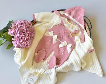 May include: A baby outfit set in a woven basket, featuring a pink romper with white bows and a matching bonnet. The set includes a cream-colored sweater and blanket. A bouquet of pink hydrangeas adds a decorative touch.