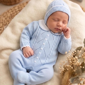 May include: A sleeping baby wearing a light blue knitted romper and matching bonnet. The romper features a cable knit design and white collar and cuffs. The bonnet has a delicate lace trim. The baby is lying on a cream-colored blanket.