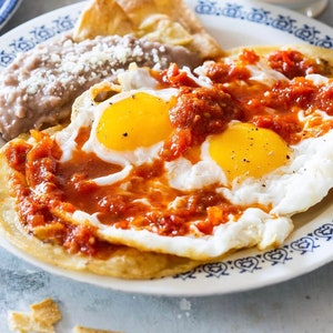 Op de afbeelding: Een bord huevos rancheros met twee gebakken eieren, rode salsa, gepureerde bonen en een knapperige tortilla chip.