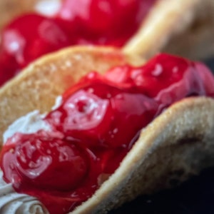 May include: A close-up of a dessert taco filled with whipped cream and topped with red cherry sauce.