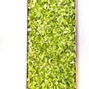 May include: A rectangular metal tray filled with chopped celery. The celery pieces are a vibrant green color and appear freshly cut. The tray is set against a white background, highlighting the bright green of the vegetables.