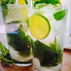 May include: Two tall clear glasses filled with a mojito cocktail containing ice cubes, fresh green mint leaves, and bright green lime slices. A clear glass straw is placed in the foreground drink. A green label with the text KING SATISFACTION is visible.