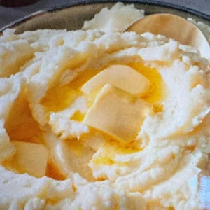 May include: A round, dark-rimmed bowl filled with creamy white mashed potatoes. Several square pats of yellow butter are placed on top, partially melted into the warm surface. A wooden spoon rests inside the bowl, and a black-handled utensil is visible.