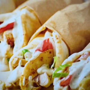 Può includere: Tre gyros di pollo avvolti in morbido pane pita e parzialmente racchiusi in carta marrone. Ogni wrap contiene pollo grigliato a fette, lattuga verde fresca, fette di pomodoro rosso e una generosa colata di salsa bianca cremosa.