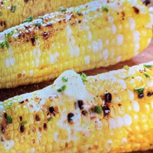 May include: Three ears of yellow corn on the cob, grilled with visible char marks. Each ear is topped with a dollop of white butter or cream and sprinkled with small, bright green chopped chives, creating a textured and colorful appearance.