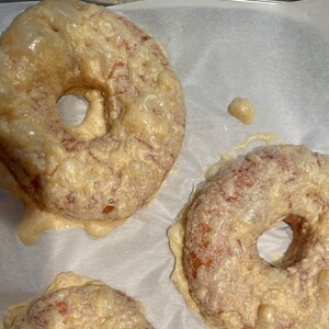 Op de afbeelding: Drie geglazuurde donuts op een bakplaat met witte bakpapierbekleding. De donuts zijn rond met een gat in het midden en hebben een lichtbruine kleur. De glazuur is wit en heeft een licht bubbelende textuur.