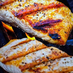 Puede incluir: Primer plano de filetes de salmón a la parrilla con marcas de parrilla. El salmón está sazonado con pimienta negra y tiene un exterior dorado con zonas rojizas. La imagen muestra el salmón cocinándose sobre una llama abierta.