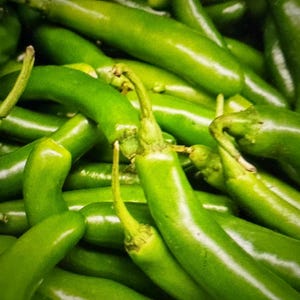 May include: A large collection of fresh, vibrant green chili peppers packed closely together. Each pepper features a smooth, glossy skin, a tapered elongated shape, and a small, light green stem attached at the top, creating a dense, uniform texture.