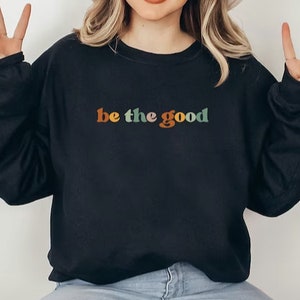 May include: Black sweatshirt featuring the phrase "be the good" in a retro font. The text is in orange, green, and brown. The sweatshirt has a crew neck design.