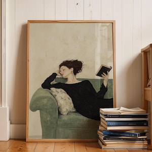 K&ouml;nnte beinhalten: Eine gerahmte Illustration einer Frau in einem schwarzen Kleid, die auf einem gr&uuml;nen Sofa liegt und ein Buch liest. Die Frau hat dunkles Haar und tr&auml;gt eine goldene Halskette. Der Hintergrund ist eine hellbeige Wand.