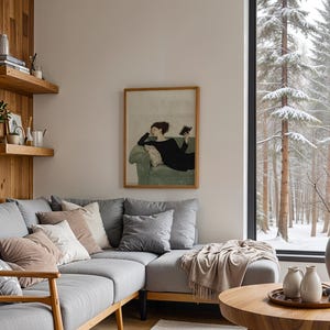 K&ouml;nnte beinhalten: Ein graues Ecksofa mit Kissen und einer Wolldecke in einem Wohnzimmer mit einem gro&szlig;en Fenster mit Blick auf einen verschneiten Wald. Ein gerahmtes Bild einer Frau, die auf einem Sofa liegt und ein Buch liest, h&auml;ngt an der Wand.