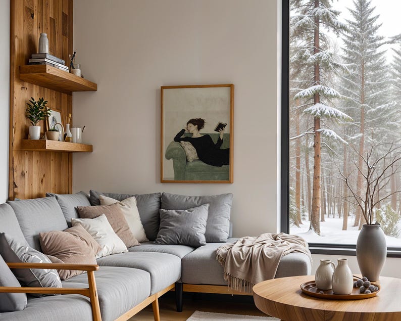 K&ouml;nnte beinhalten: Ein graues Ecksofa mit Kissen und einer Wolldecke in einem Wohnzimmer mit einem gro&szlig;en Fenster mit Blick auf einen verschneiten Wald. Ein gerahmtes Bild einer Frau, die auf einem Sofa liegt und ein Buch liest, h&auml;ngt an der Wand.