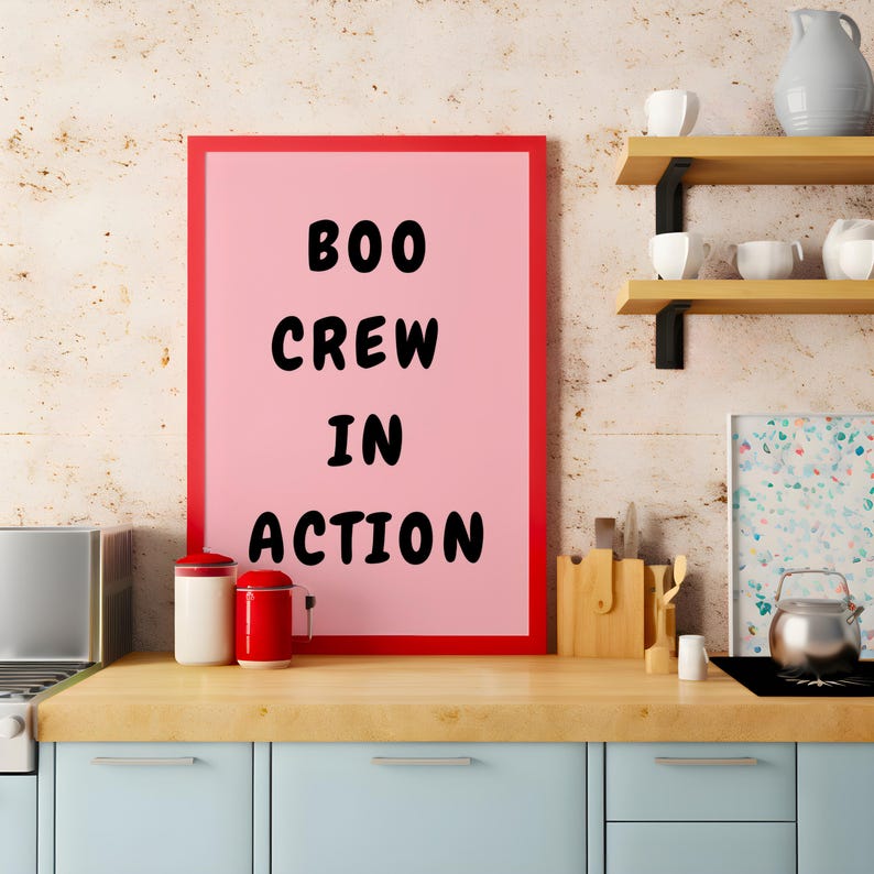 Puede incluir: Un p&oacute;ster enmarcado rosa con el texto "BOO CREW IN ACTION" en letras negras. El p&oacute;ster est&aacute; colgado en una pared en un ambiente de cocina.
