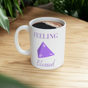 May include: White ceramic mug with a purple four-sided die graphic and the text "Feeling Blessed". The mug is filled with coffee.