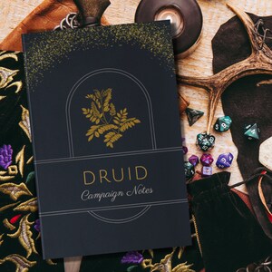May include: A dark blue notebook with gold lettering that says "DRUID Campaign Notes". The cover features a gold illustration of a plant with a gold arch behind it. The notebook is surrounded by dice, a leather pouch, and other fantasy-themed items.