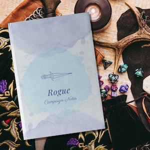 Puede incluir: Un cuaderno azul y blanco con un fondo de acuarela y un dibujo lineal de una daga. El cuaderno dice "Rogue Campaign Notes" en la portada.