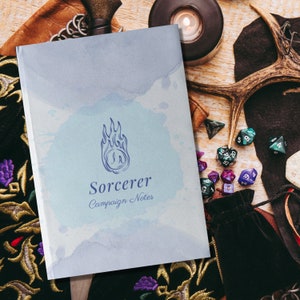 Puede incluir: Un cuaderno con un diseño de acuarela azul y las palabras "Sorcerer Campaign Notes". El cuaderno presenta un logotipo de llama. Dados, una vela y otros accesorios de juegos de fantasía rodean el cuaderno.