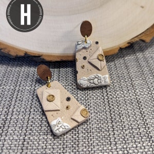May include: A pair of beige and white owl-themed earrings with gold accents. The earrings are shaped like rectangles and feature a design of owls, envelopes, and a brown background.