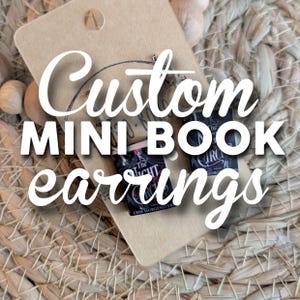 May include: Miniature book earrings with the text "The Night Circus" and "Custom MINI BOOK earrings" on a brown card. The earrings are displayed on a woven background.