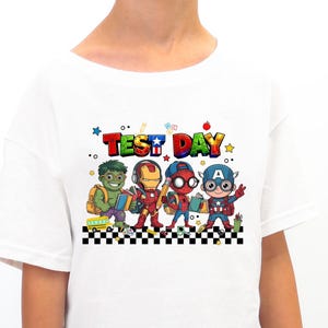 Puede incluir: Camiseta blanca con un gráfico colorido de superhéroes de dibujos animados, incluyendo a Hulk, Iron Man, Spider-Man y Capitán América. El texto "TEST DAY" está sobre los personajes. El diseño está sobre un patrón de cuadros.