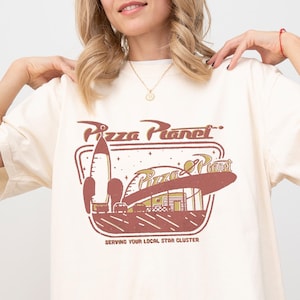 Puede incluir: Camiseta color crema con un gr&aacute;fico de Pizza Planet. El dise&ntilde;o incluye un cohete, un restaurante y el texto "Pizza Planet" y "Serving Your Local Star Cluster" en marr&oacute;n. La camiseta tiene mangas cortas y un corte holgado.