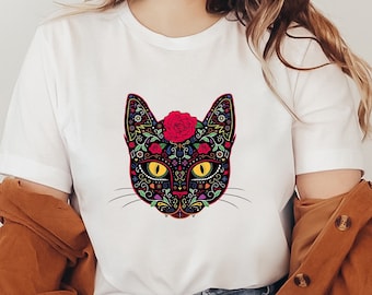 May include: White t-shirt featuring a colorful cat face design. The cat's face is black with floral and decorative patterns, yellow eyes, and a red rose on its head. The shirt is worn by a person wearing a brown hat and a brown jacket.
