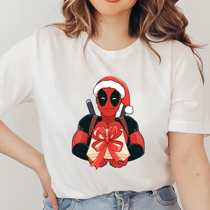 Pode incluir: Camiseta branca com um personagem de desenho animado Deadpool usando um gorro de Papai Noel e segurando um presente embrulhado com um laço vermelho. O gráfico é nas cores vermelho, preto e bege. A camiseta é de estilo casual.
