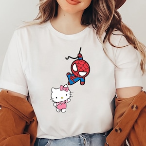 May include: White t-shirt featuring a cartoon graphic of Spider-Man and Hello Kitty. Spider-Man is depicted in his classic red and blue suit, while Hello Kitty is wearing a pink dress and bow. The word "White" is printed in the upper right corner.