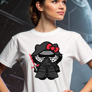 Puede incluir: Camiseta blanca con una combinación de dibujos animados de Kylo Ren y Hello Kitty. El personaje lleva un casco negro con un lazo rojo y sostiene un sable de luz rojo. El fondo muestra una escena espacial.