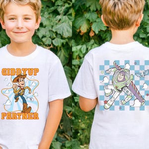 May include: White t-shirt featuring a cartoon graphic of Woody from Toy Story with the text "Giddyup Partner". The back of the shirt has a graphic of Buzz Lightyear. The shirt is worn by a young child.