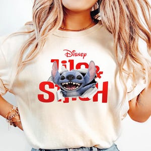 Puede incluir: Camiseta color crema con el logo de Disney y las palabras "Lilo & Stitch" en rojo. El dise&ntilde;o incluye al personaje azul Stitch saliendo del texto, sonriendo. La camiseta se combina con vaqueros azules.