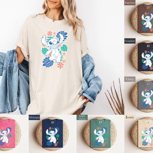 Puede incluir: Una camiseta color crema con un personaje de dibujos animados con hojas y flores tropicales azules y verdes. La camiseta se muestra en una persona. Tambi&eacute;n se muestran camisetas adicionales en varios colores, incluyendo espresso, grafito y ar&aacute;ndano.