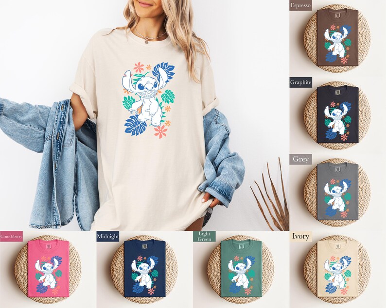 Puede incluir: Una camiseta color crema con un personaje de dibujos animados con hojas y flores tropicales azules y verdes. La camiseta se muestra en una persona. Tambi&eacute;n se muestran camisetas adicionales en varios colores, incluyendo espresso, grafito y ar&aacute;ndano.