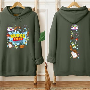 K&ouml;nnte beinhalten: Ein olivgr&uuml;ner Kapuzenpullover mit einem Front-Grafik-Aufdruck &bdquo;NURSE STORY&ldquo; und medizinischen Illustrationen. Die R&uuml;ckseite zeigt ein vertikales Design mit medizinischen Symbolen. Der Hoodie hat eine Kapuze mit Kordelzug und eine Vordertasche.