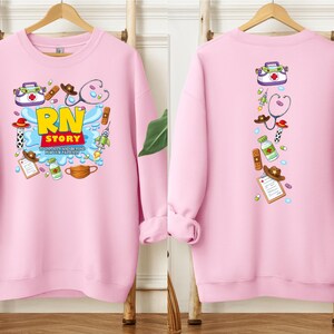 K&ouml;nnte beinhalten: Ein hellrosa Sweatshirt mit einem farbenfrohen Grafikdesign auf der Vorder- und R&uuml;ckseite. Die Grafik auf der Vorderseite zeigt "RN STORY" mit medizinischen Illustrationen. Die R&uuml;ckseite zeigt eine vertikale Anordnung &auml;hnlicher medizinischer Illustrationen.