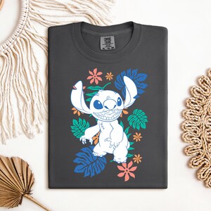 Puede incluir: Camiseta gris oscuro con un personaje de dibujos animados blanco con orejas grandes y una amplia sonrisa, rodeado de dise&ntilde;os florales y de hojas coloridas. La camiseta est&aacute; doblada sobre una superficie blanca, mostrando un dise&ntilde;o juguet&oacute;n y vibrante.