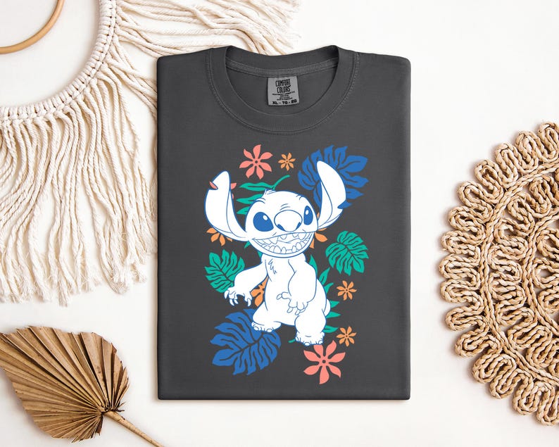 Puede incluir: Camiseta gris oscuro con un personaje de dibujos animados blanco con orejas grandes y una amplia sonrisa, rodeado de dise&ntilde;os florales y de hojas coloridas. La camiseta est&aacute; doblada sobre una superficie blanca, mostrando un dise&ntilde;o juguet&oacute;n y vibrante.