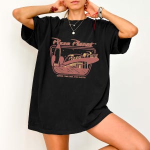 Puede incluir: Camiseta negra con un gr&aacute;fico de Pizza Planet. El dise&ntilde;o incluye un cohete, un restaurante y el texto "Pizza Planet" en un estilo retro. Tambi&eacute;n est&aacute; presente la frase "Serving Your Local Star Cluster".