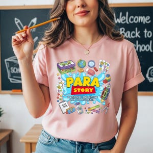 K&ouml;nnte beinhalten: Hellrosa T-Shirt mit dem farbenfrohen Schriftzug "PARA STORY". Das Design zeigt schulische Illustrationen wie einen Globus, einen Radiergummi und eine Rakete. Der Text "TO INFINITY AND BEYOND FOR OUR STUDENTS" ist ebenfalls enthalten.
