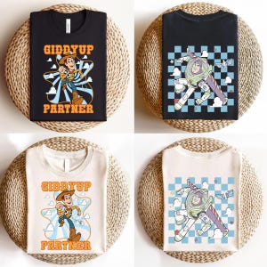 May include: Four t-shirts with cartoon graphics. Two black t-shirts and two off-white t-shirts. One black t-shirt has a graphic of Buzz Lightyear. The other black t-shirt has a graphic of Woody with the text "Giddyup Partner". The other two shirts have the same graphics.