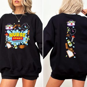 K&ouml;nnte beinhalten: Schwarzes Sweatshirt mit wei&szlig;em Kragen und einem farbenfrohen "Nurse Story"-Motiv auf der Vorderseite. Das Design umfasst medizinische Werkzeuge, einen Cowboyhut und den Text "To infinity and beyond for our patients."