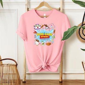 May include: A light pink t-shirt with a graphic design featuring medical-themed illustrations and the text "Peds Care Story". The design includes a stethoscope, bandages, and a face mask. The shirt is hanging on a wooden rack.