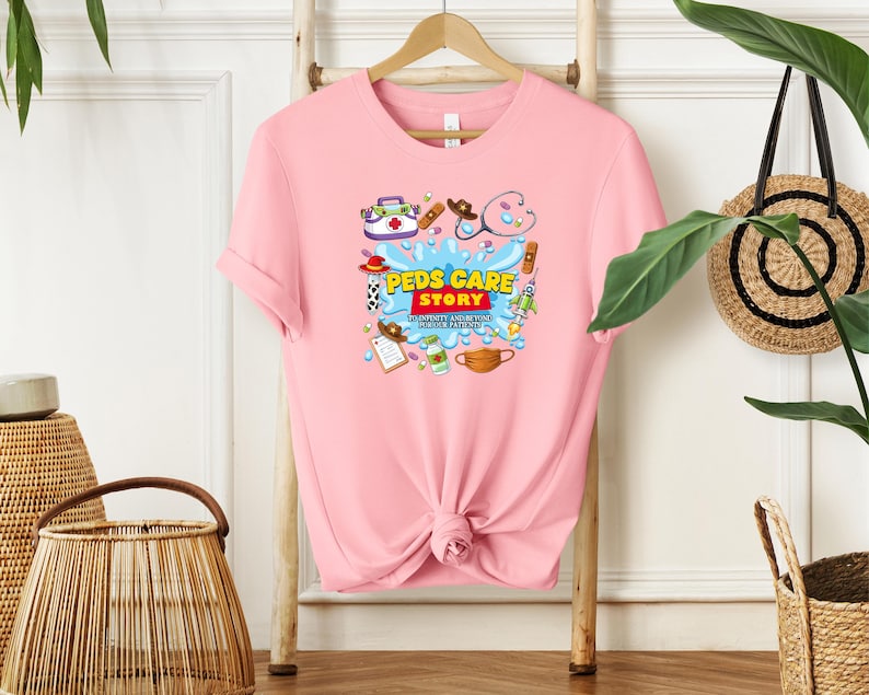May include: A light pink t-shirt with a graphic design featuring medical-themed illustrations and the text "Peds Care Story". The design includes a stethoscope, bandages, and a face mask. The shirt is hanging on a wooden rack.