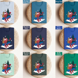 Puede incluir: Una colecci&oacute;n de camisetas en varios colores, cada una con un gr&aacute;fico de un superh&eacute;roe con gorro de Pap&aacute; Noel, llevando un saco de regalos y un &aacute;rbol de Navidad. Los colores incluyen azul claro, marino, verde azulado y verde.