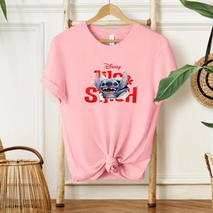Puede incluir: Camiseta rosa claro con un dise&ntilde;o de Stitch de Disney. El dise&ntilde;o incluye el logotipo de Disney y las palabras "Lilo & Stitch" en rojo. La camiseta est&aacute; colgada en un perchero de madera.