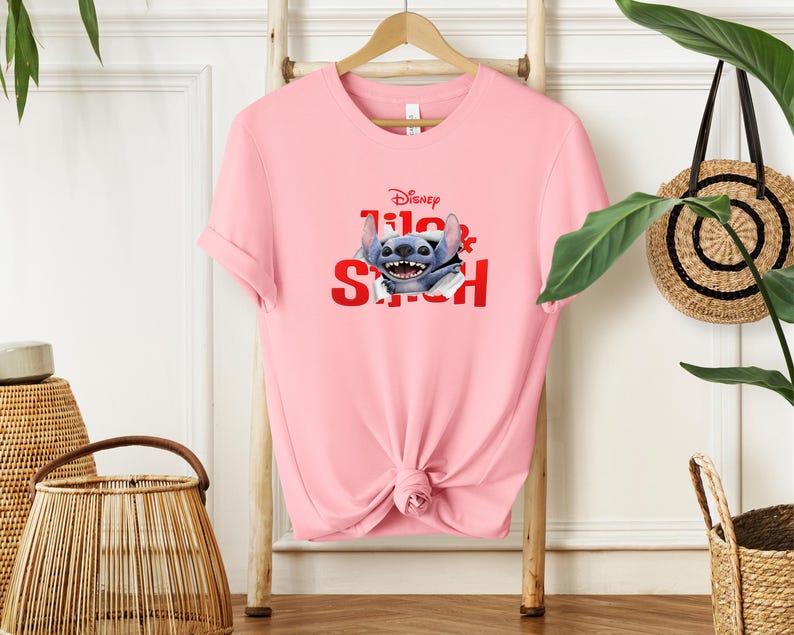 Puede incluir: Camiseta rosa claro con un dise&ntilde;o de Stitch de Disney. El dise&ntilde;o incluye el logotipo de Disney y las palabras "Lilo & Stitch" en rojo. La camiseta est&aacute; colgada en un perchero de madera.